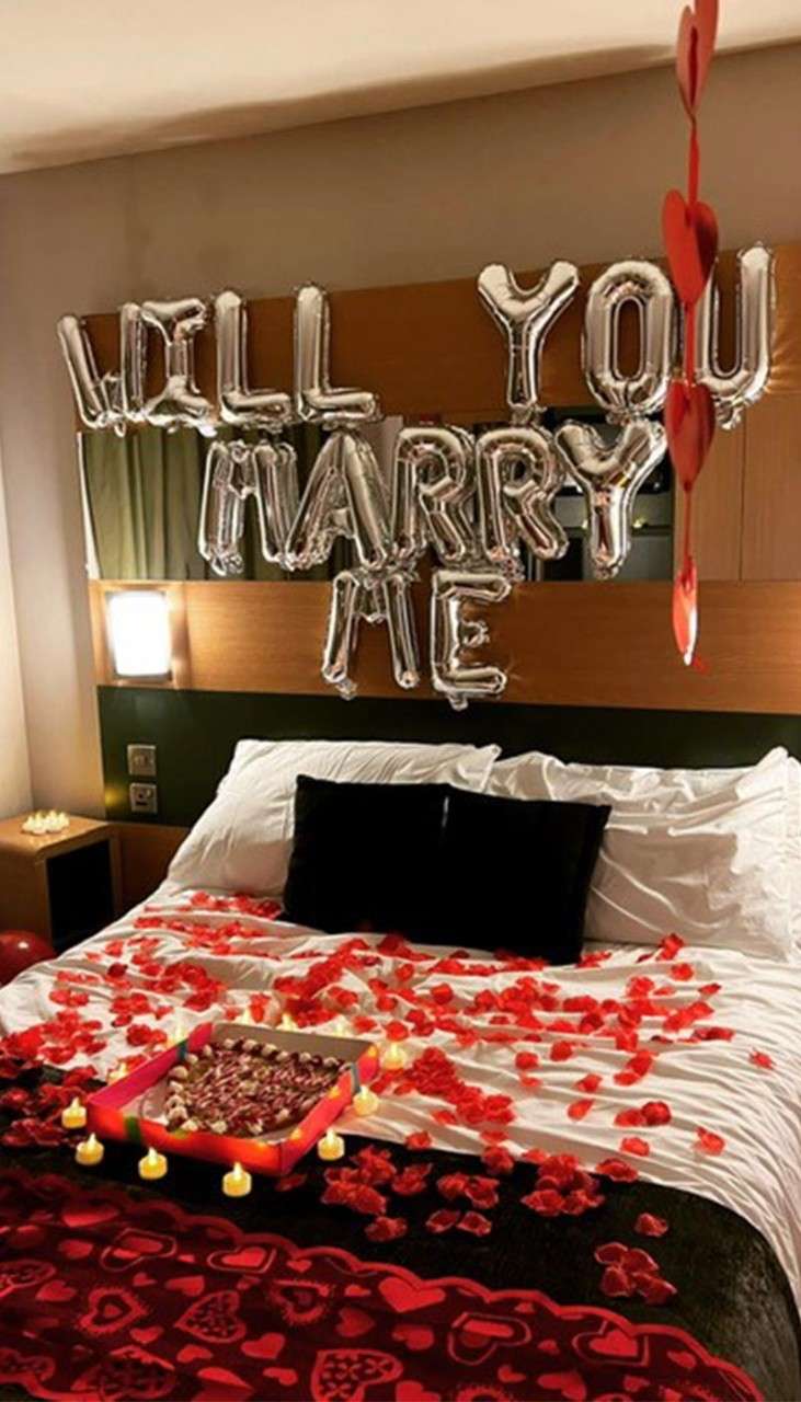 "Will you marry me" balloons above a bed with rose petals on displayed by a guest.