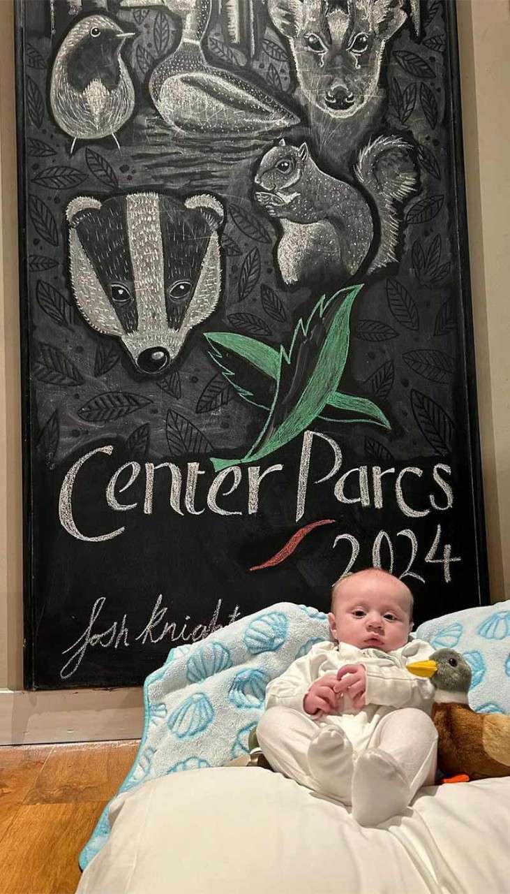 A baby layed in front of a blackboard in a lodge with a personalised drawing out of chalk.